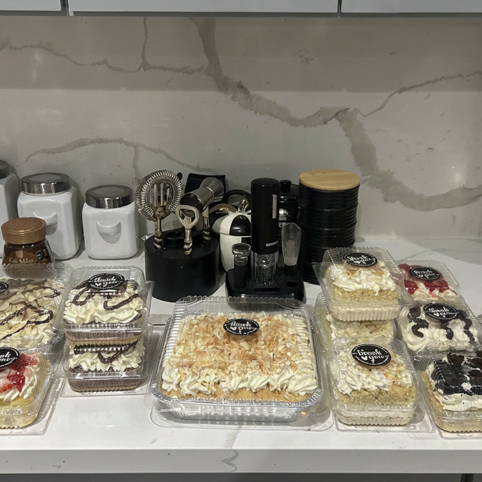 Counter displays assorted packaged cakes and layered desserts beside coffee equipment and storage canisters.